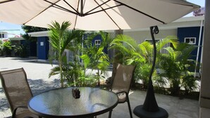 Un patio al aire libre con una mesa redonda y sillas, un paraguas grande y diversas plantas tropicales.