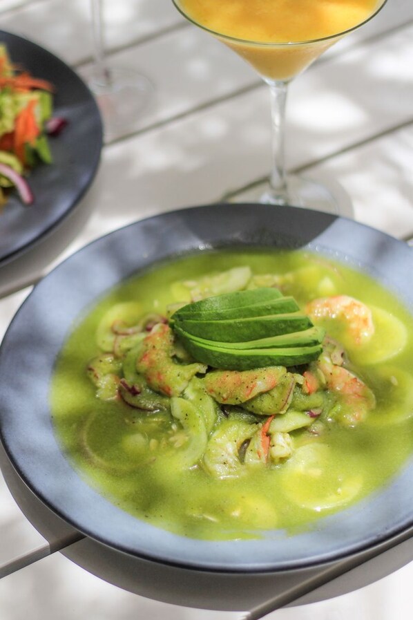 A bowl of green soup with shrimp and avocado garnish, a glass of orange juice, and a plate of salad in the background.