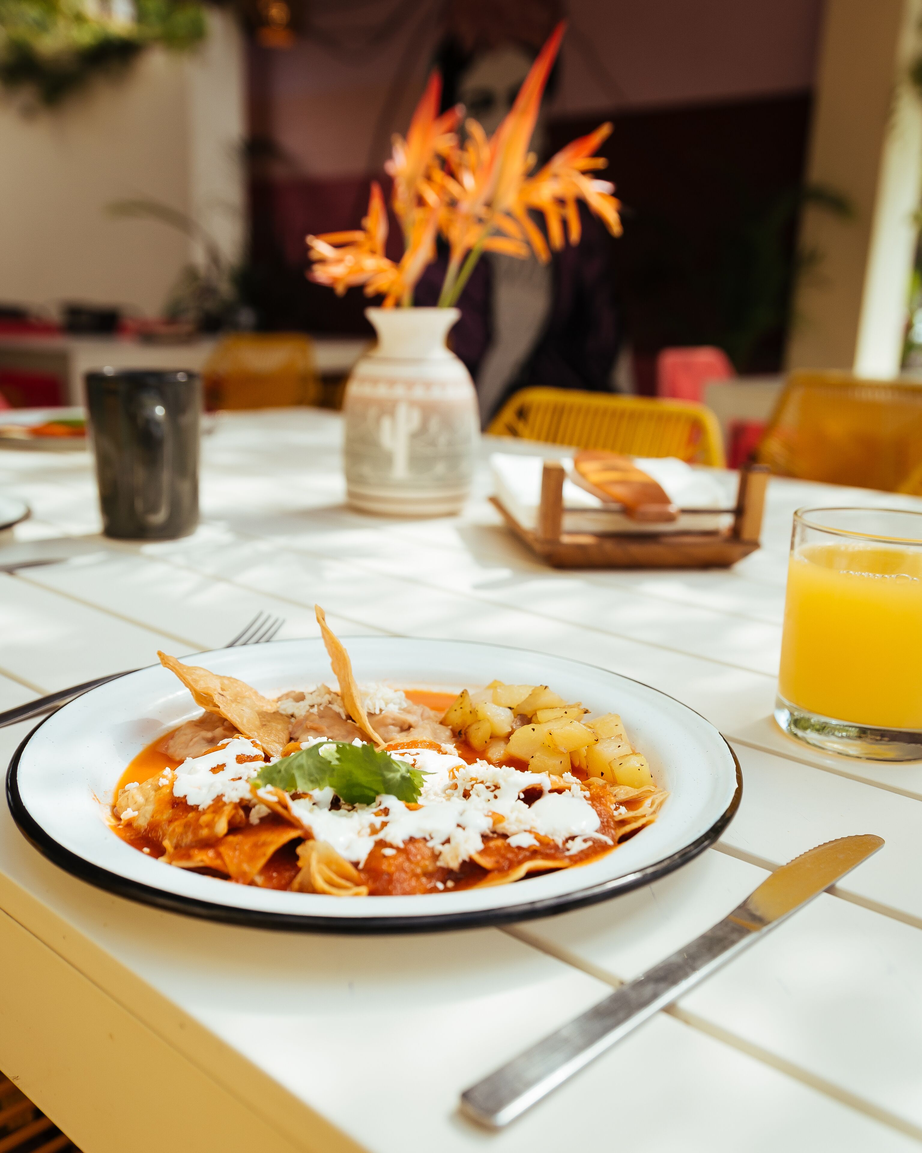 Una mesa de comedor con un plato de enchiladas, un vaso de jugo de naranja y un florero con flores.