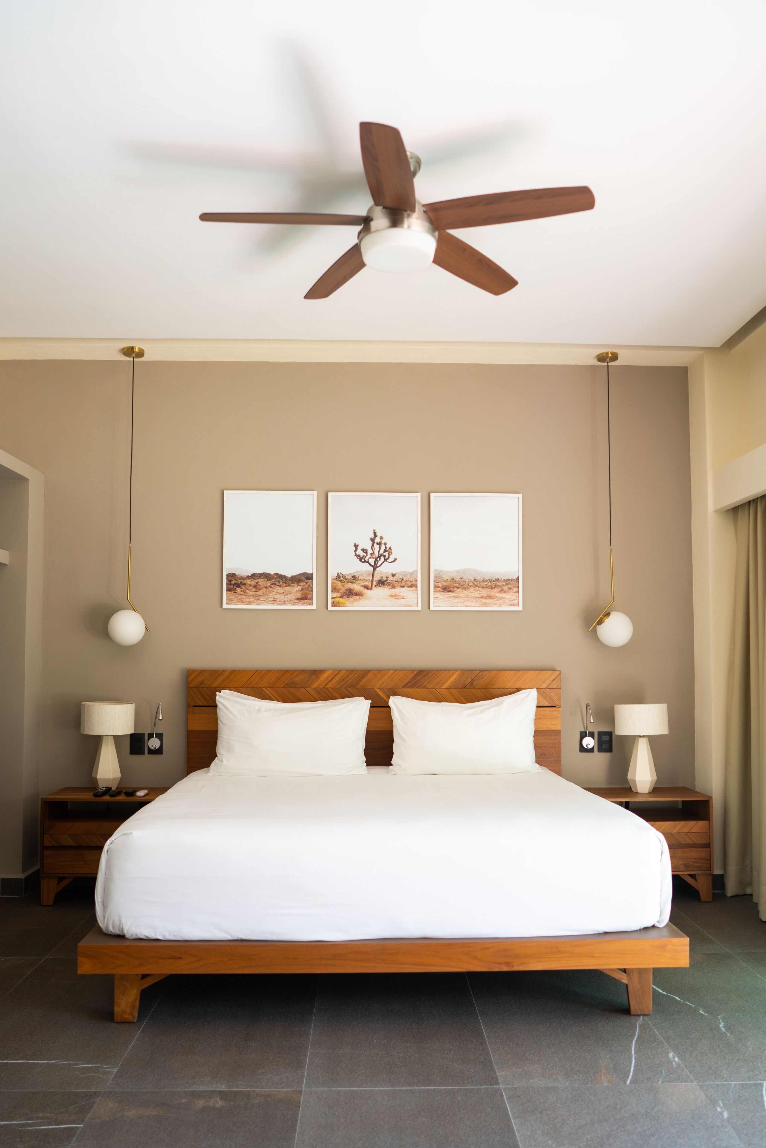 A bedroom with a wooden bed, two bedside tables with lamps, a ceiling fan, and three framed pictures on the wall.