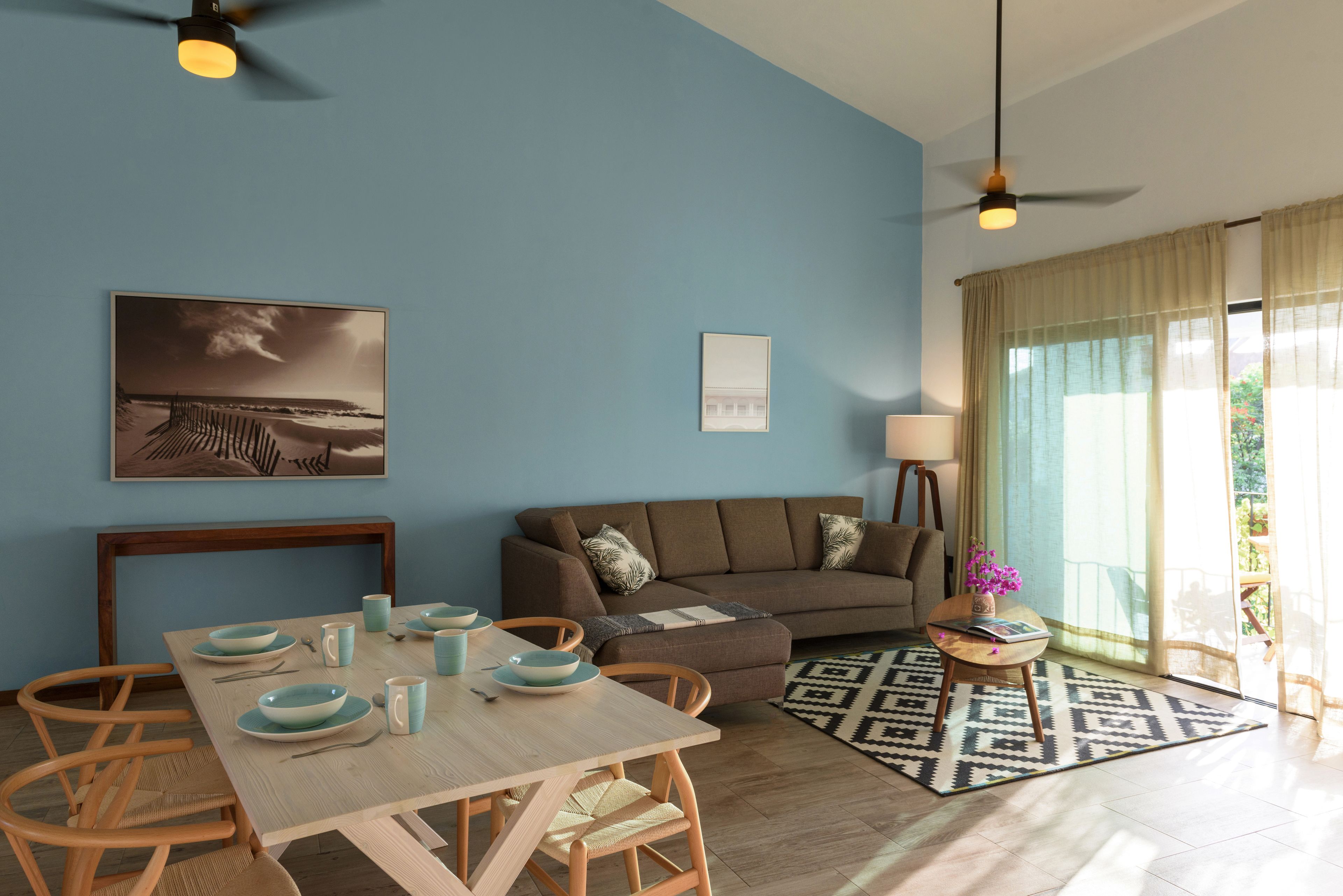 A modern living room with a dining area, featuring a blue wall, a sofa, a coffee table, and a view of the outdoors.