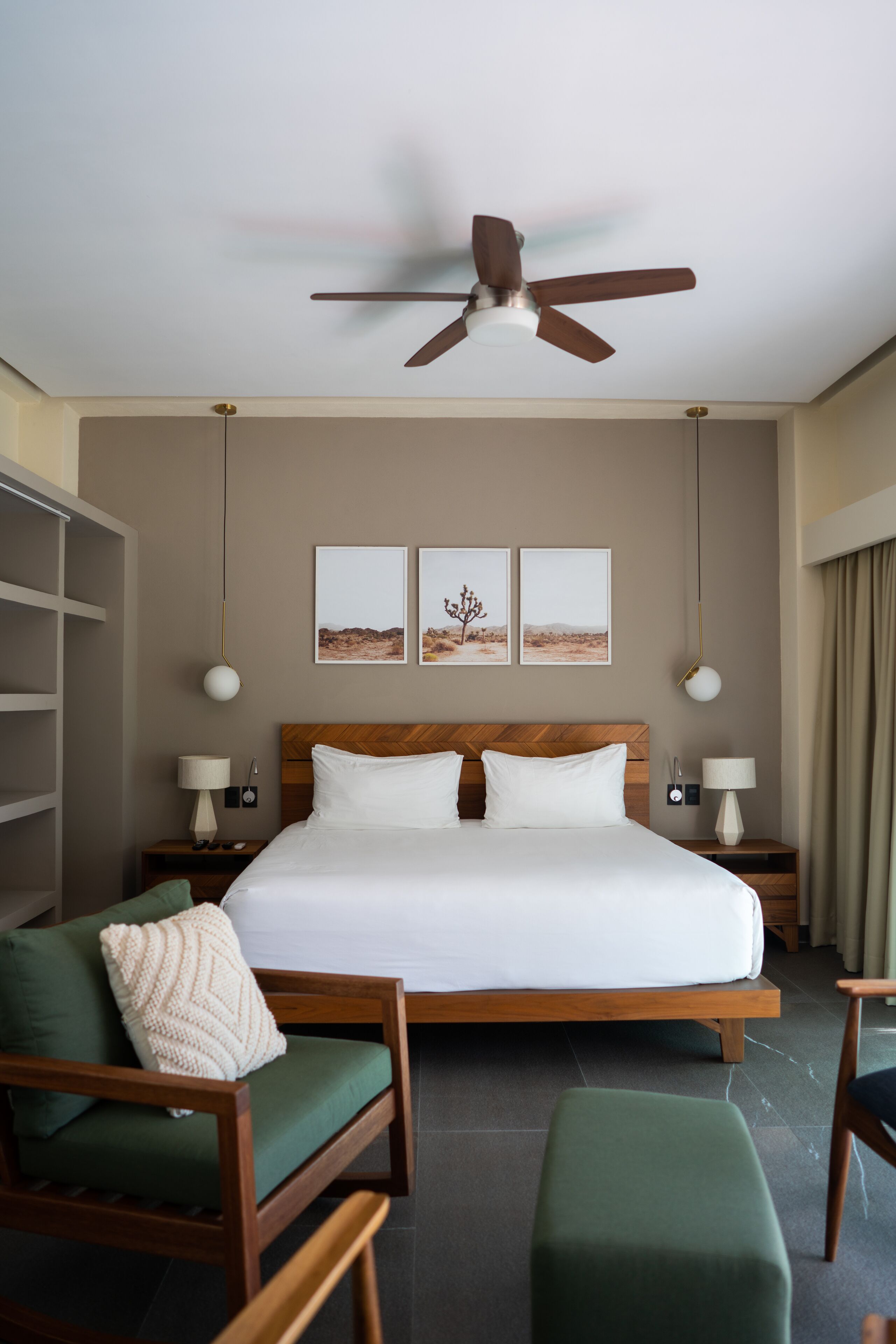 Una habitación de hotel con una cama grande, cabecera de madera, dos mesitas de noche con lámparas, un ventilador de techo, un sillón verde y un reposapiés verde.