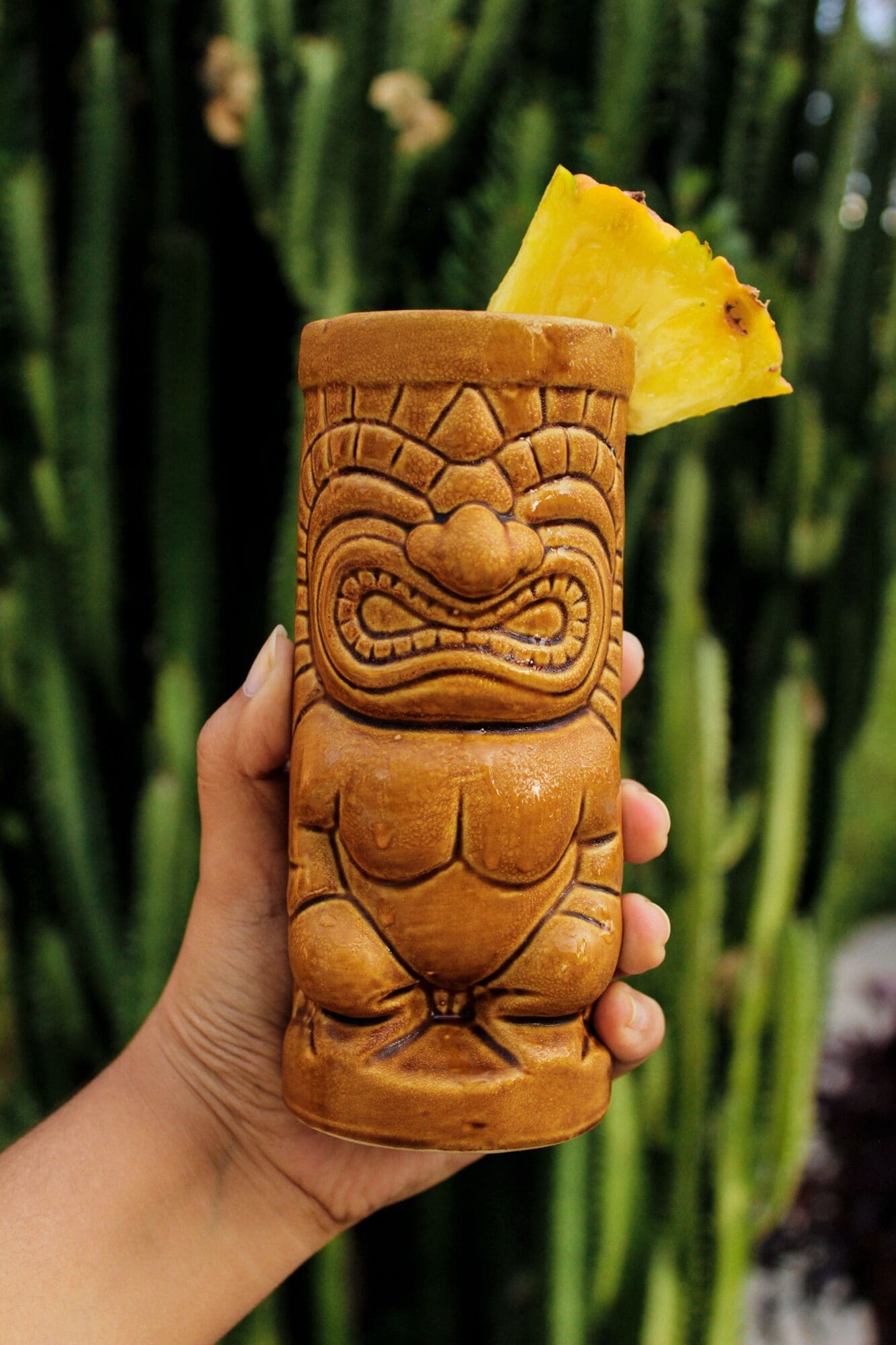Eine Hand hält einen Tiki-Becher mit einem geschnitzten Gesicht und einer Ananas darauf.