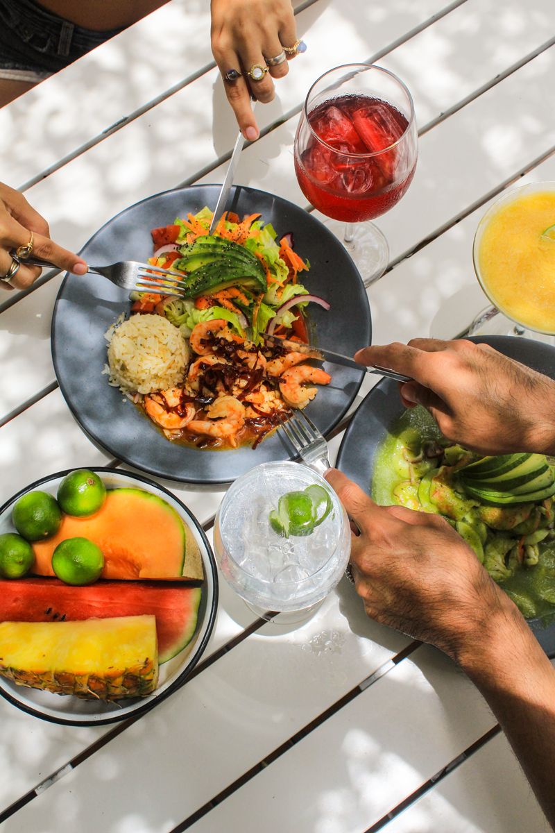 A table with a meal consisting of rice, shrimp, vegetables, and a glass of red drink. There are also bowls of fruit and a glass of clear drink with ice.