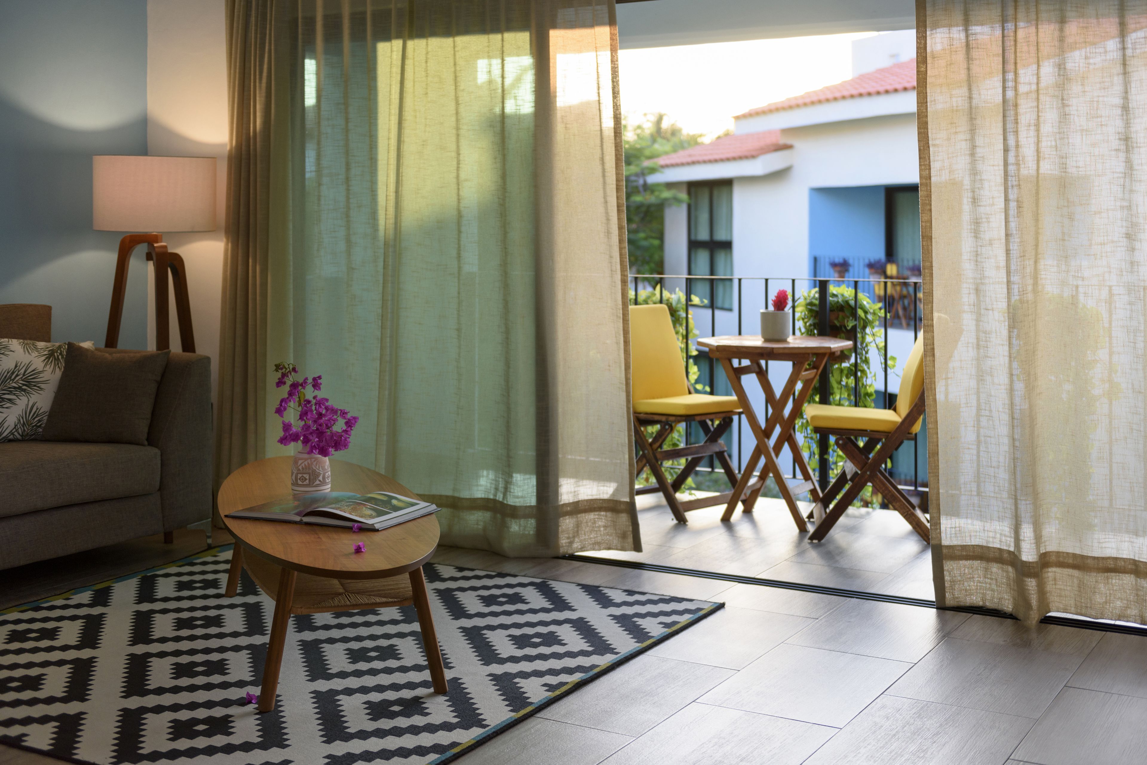 Ein Wohnzimmer mit einer Couch, einem Couchtisch mit Buch und Blumen, einem gemusterten Teppich und Blick auf einen Balkon mit Stühlen und Pflanzen.