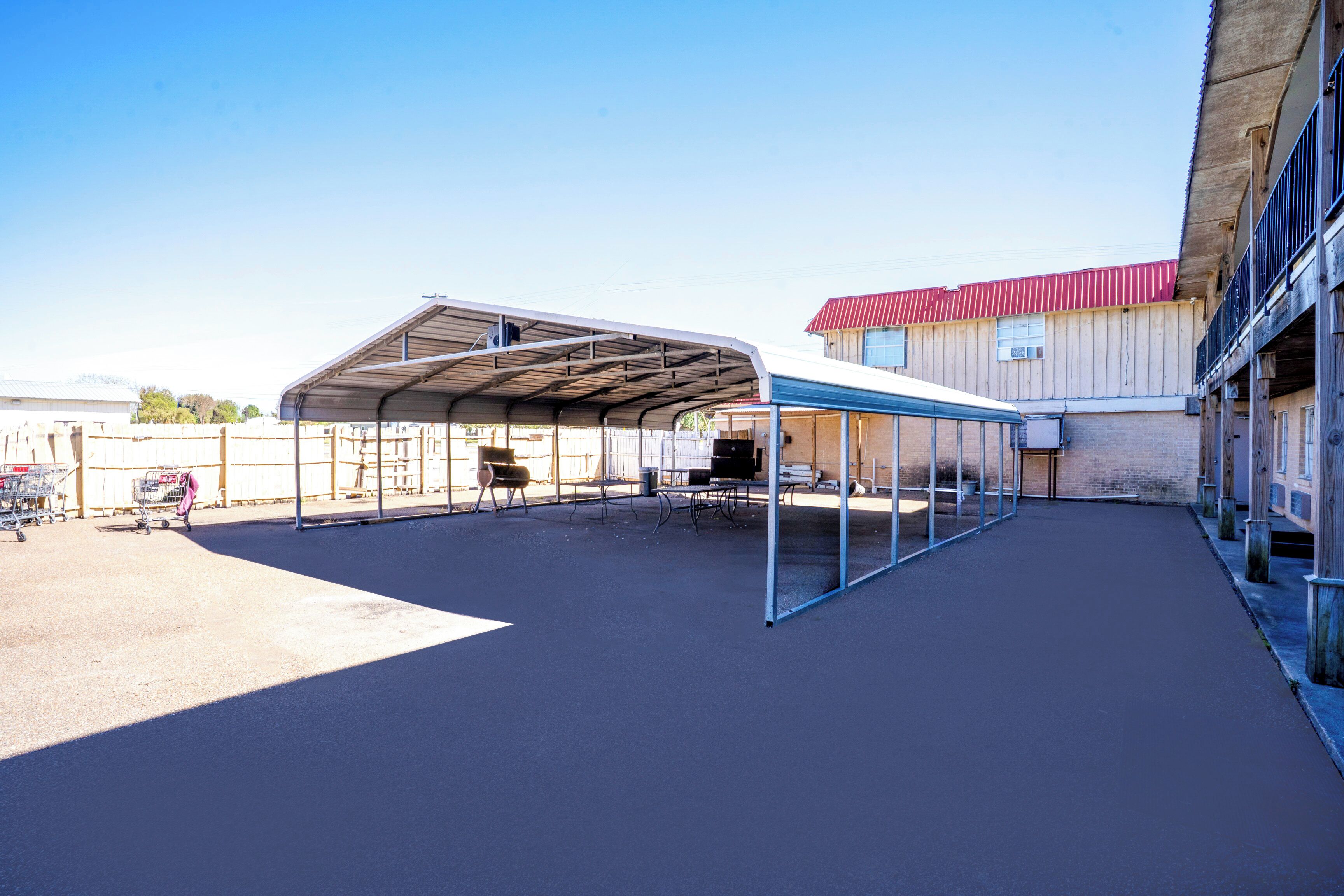 An outdoor covered area with tables and chairs, a building with a red roof, and a fenced perimeter.