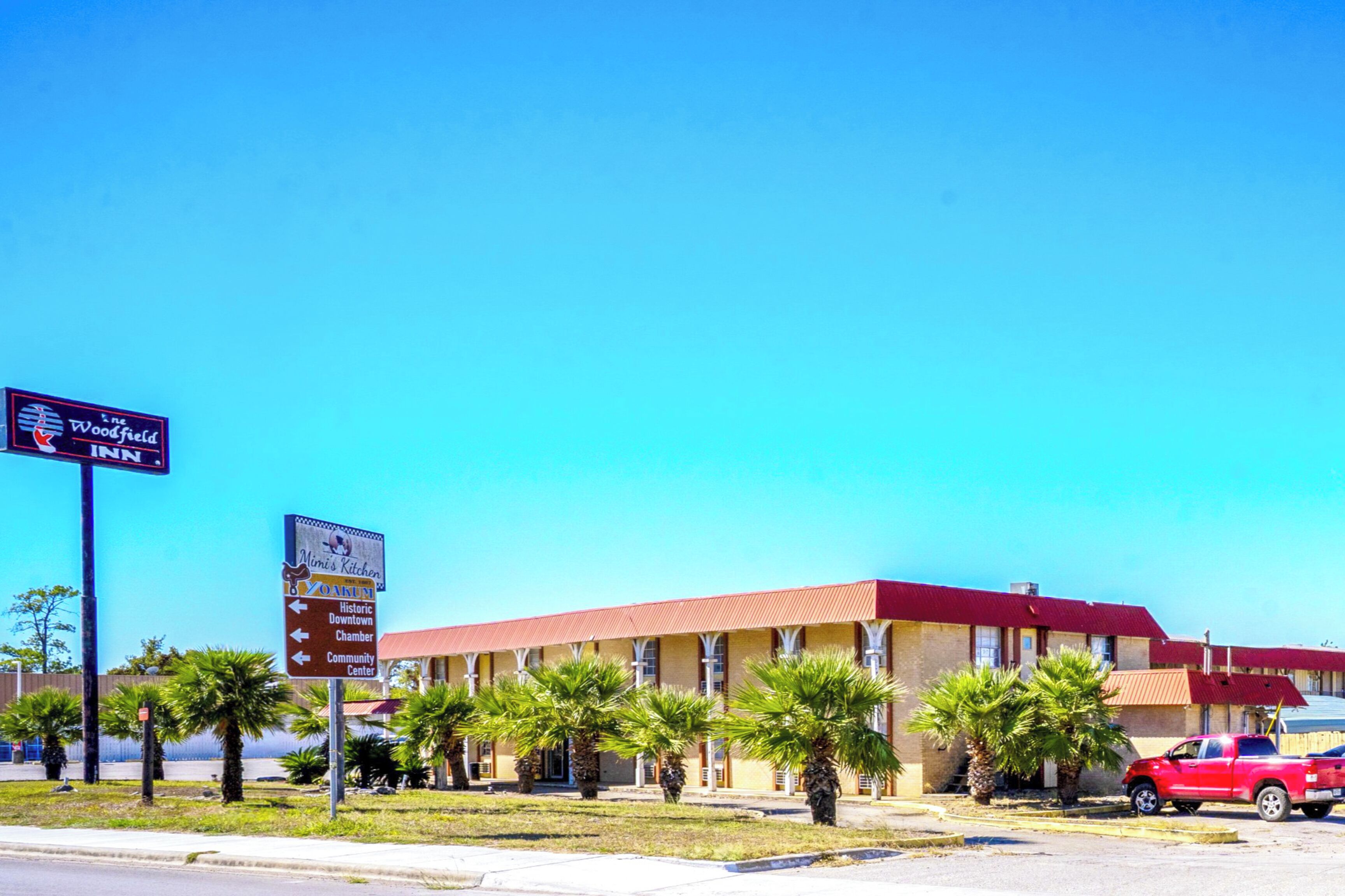 Ein Motel mit rotem Dach und Palmen davor. Ein Schild weist auf das Woodfield Inn hin, ein weiteres auf Mini's Kitchen.