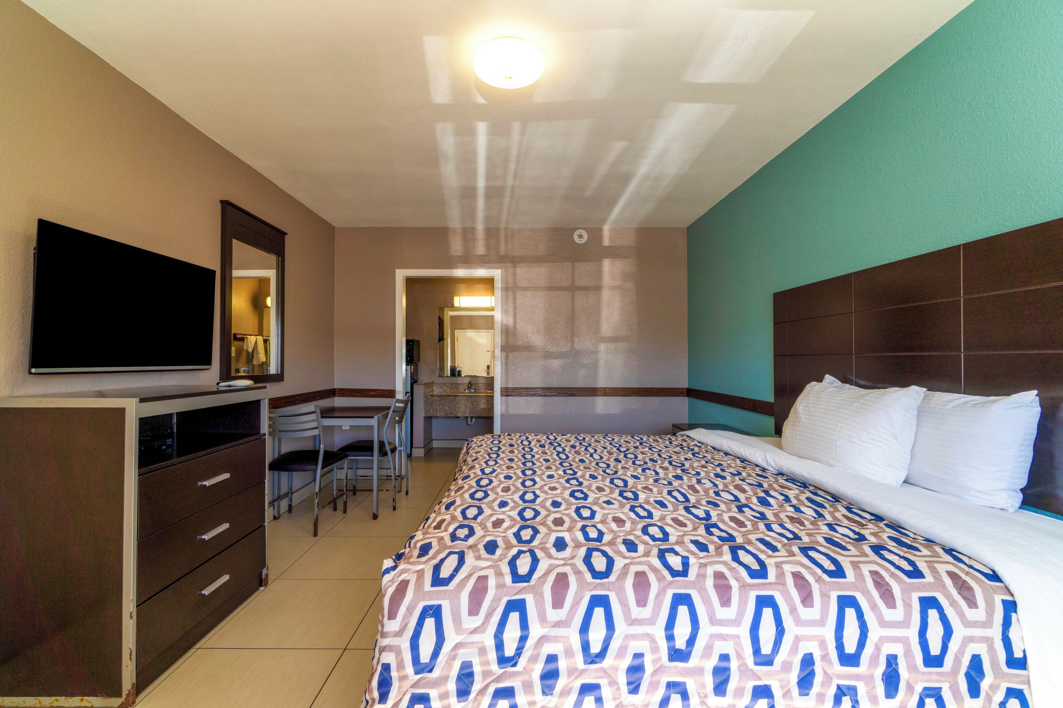 A hotel room with a bed, a TV mounted on a cabinet, a desk with a chair, and a bathroom visible through an open door.