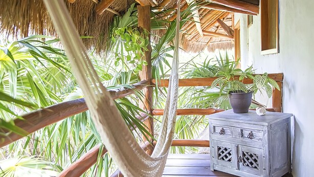 A hammock hanging between wooden beams, a potted plant on a wooden side table, and a thatched roof structure. - Holbox Deluxe Wellness Apartments (Isla Holbox)