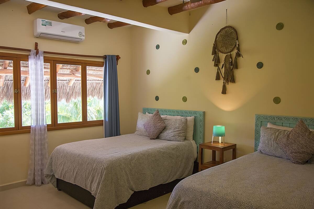 A room with two beds, a wall-mounted air conditioner, a dreamcatcher, and a window with curtains.