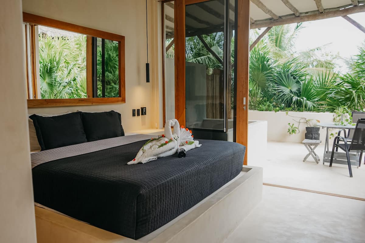 A bedroom with a large bed, a window with a view of greenery, and a sliding glass door leading to an outdoor area with a table and chairs.