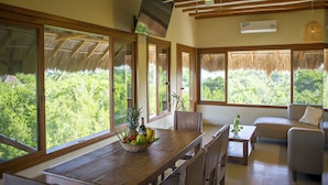 A spacious room with a wooden dining table, a sofa, and a TV mounted on the wall. Large windows offer a view of lush greenery outside. - Holbox Deluxe Wellness Apartments (Isla Holbox)