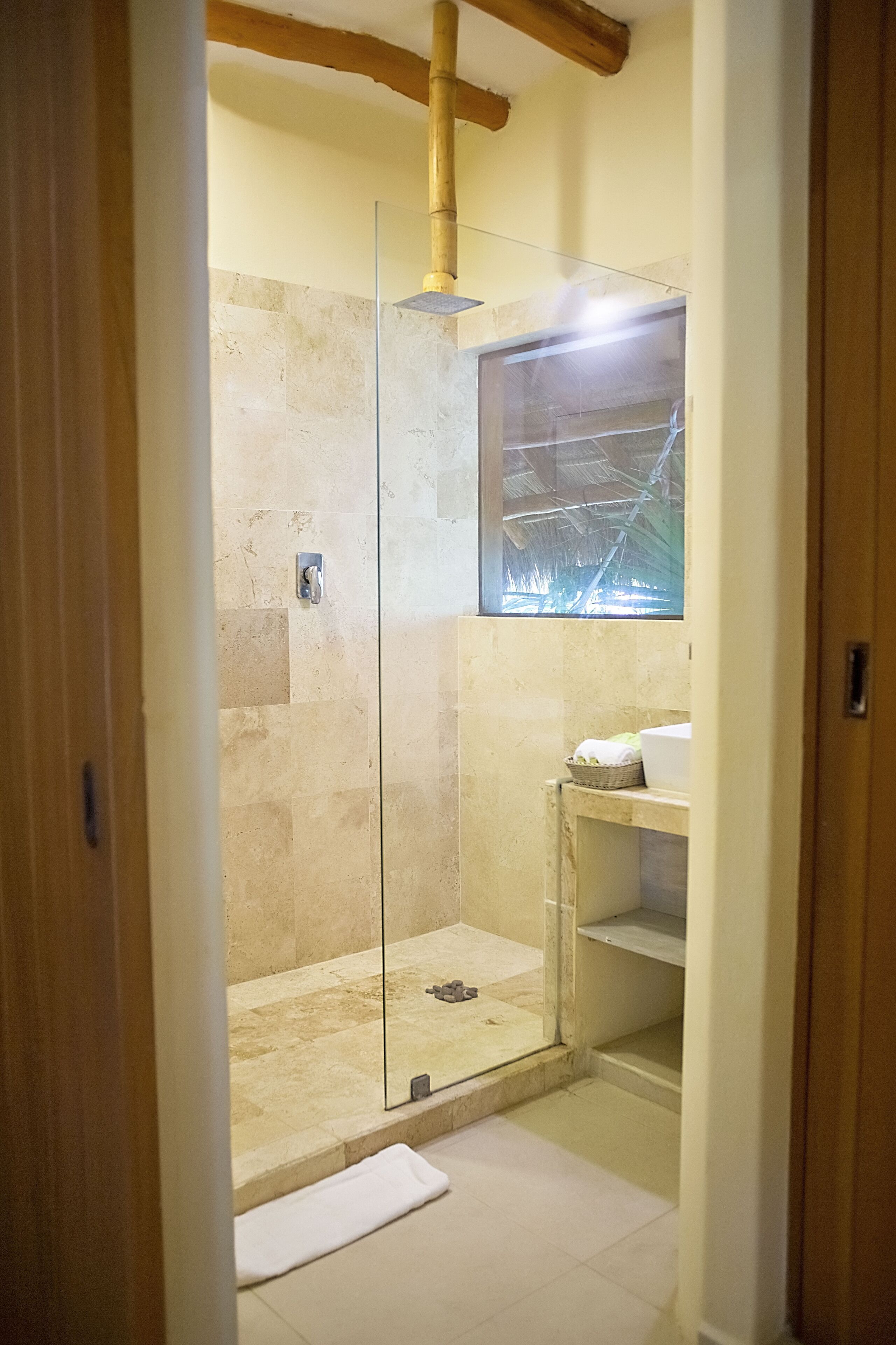 Ducha con cerramiento de vidrio, azulejos color beige, viga de techo de madera y un estante pequeño con una toalla blanca.
