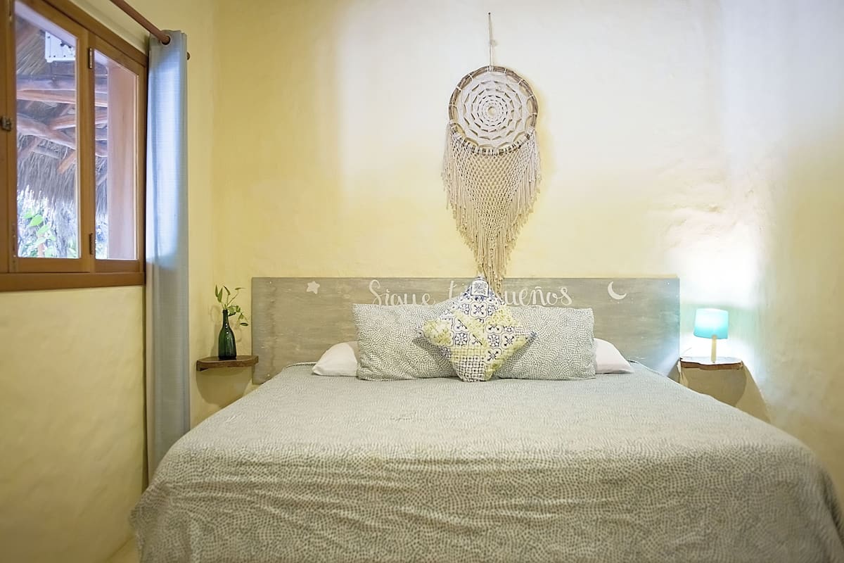A bedroom with a bed, a dreamcatcher, a wall with a sign, a window, and a nightstand with a lamp.