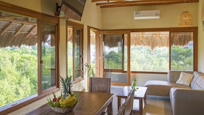 A room with a wooden ceiling, large windows with thatched roofs, a wooden table with a fruit bowl, a sofa, and a hanging light fixture. - Holbox Deluxe Wellness Apartments (Isla Holbox)