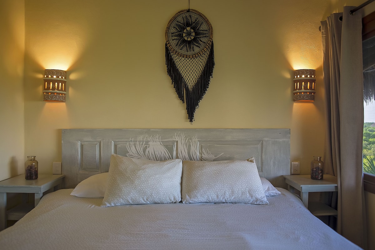 A bedroom with a bed, two bedside tables, a wall-mounted decorative item, and a window with curtains.