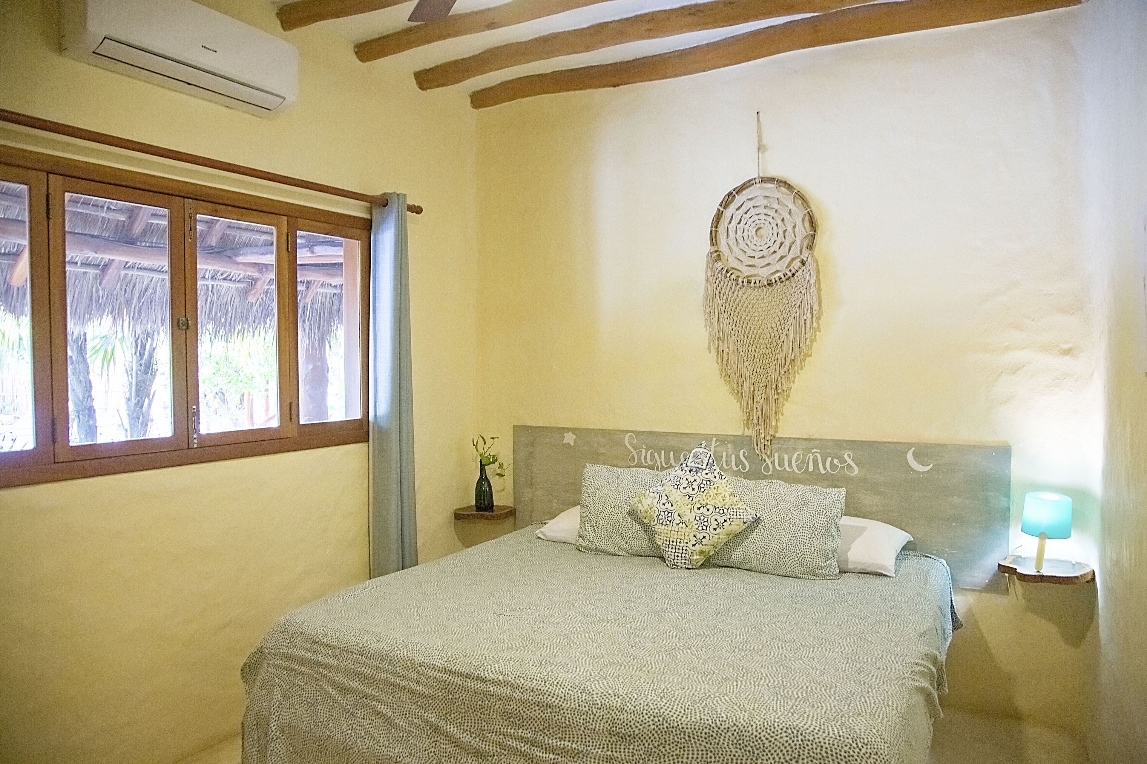 Un dormitorio con cama, un atrapasueños, una lámpara de noche y una ventana con vistas a árboles.