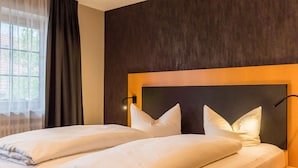 Una cama doble con cabecera oscura y dos mesitas de noche, una ventana con cortinas ligeras y vistas a la vegetación.