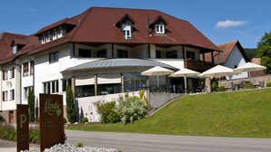 Un hotel con techo de tejas rojas, una zona de estar al aire libre cubierta con sombrillas y un cartel que indica que es un hotel 'Linde'.