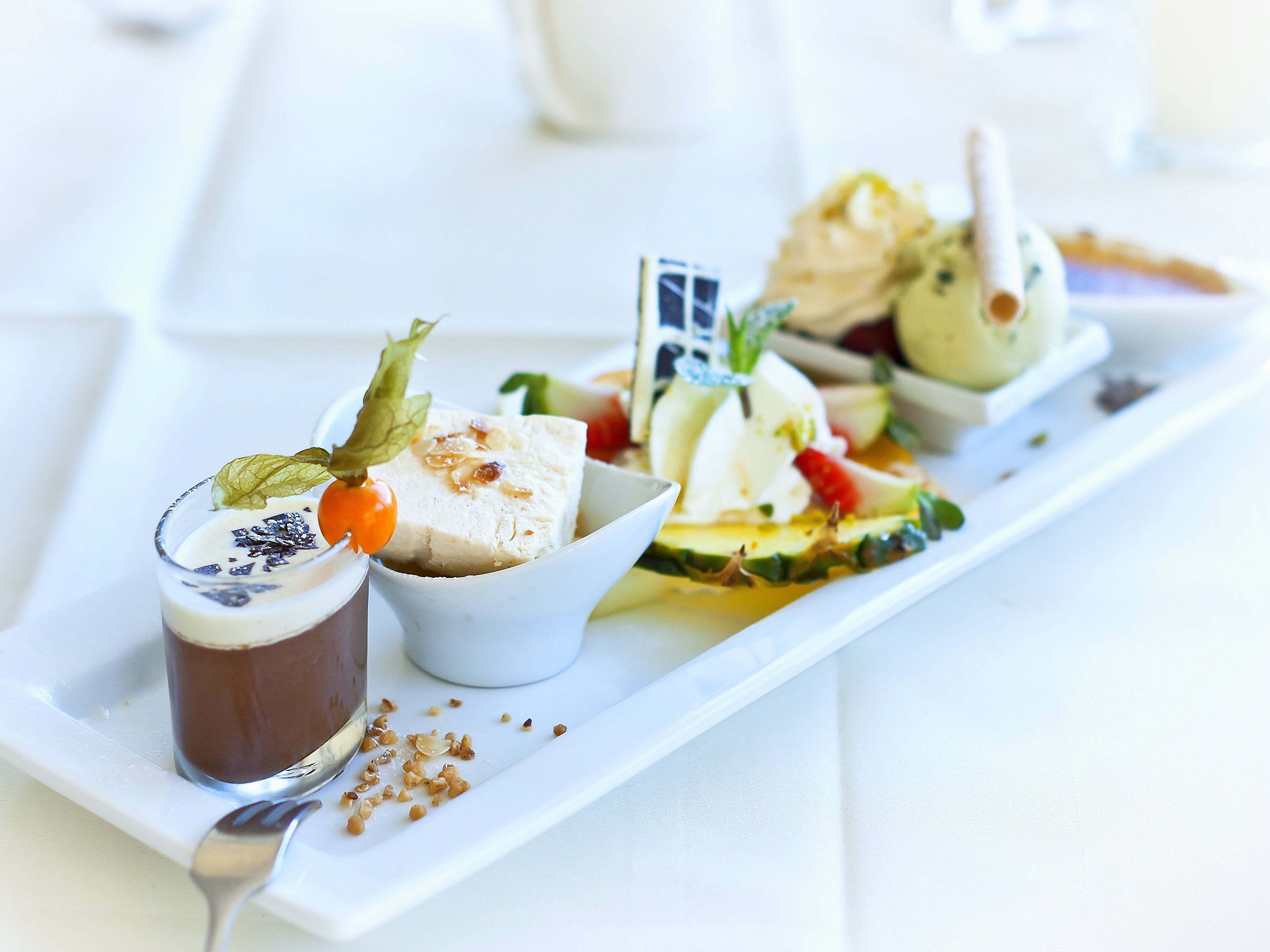 A white rectangular plate with various food items including a dessert with a glass of chocolate, a white creamy dessert with raisins, and a salad with vegetables and a decorative item.