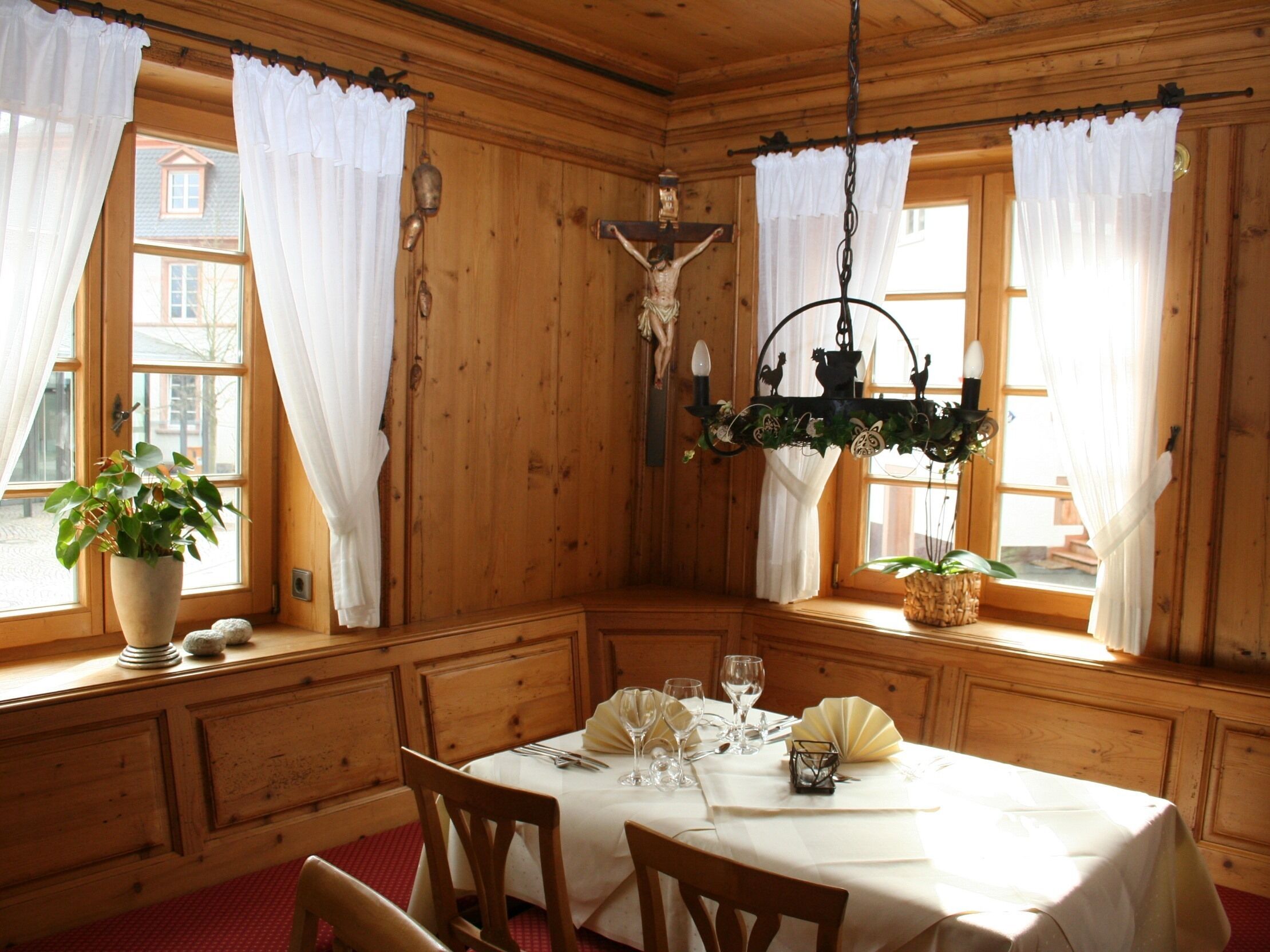 A wooden interior with a dining table set for two, a crucifix on the wall, and a hanging light fixture.