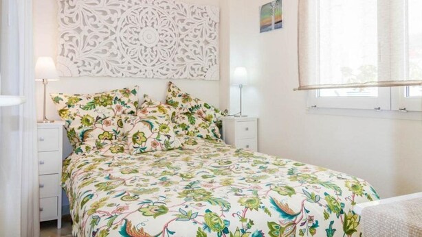 Un dormitorio con colcha estampada, un panel decorativo en la pared, una mesita de noche con lámpara y una ventana con vistas.