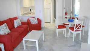 Una sala de estar moderna con un sofá rojo, una mesa de centro blanca y sillas a juego. Hay un comedor con una mesa y sillas blancas, y una cocina visible a través de un arco.
