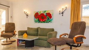 Una sala de estar con un sofá verde, dos sillones marrones, una mesa de centro de vidrio y un cuadro de flores rojas en la pared.
