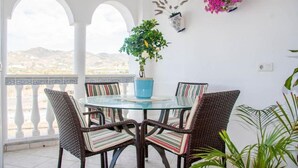 Mesa de vidrio con sillas de mimbre, planta en maceta y arreglo floral colgante en una habitación luminosa con vistas.