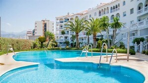 Una piscina de borde curvo, rodeada de jardines con palmeras y un edificio con balcones al fondo.