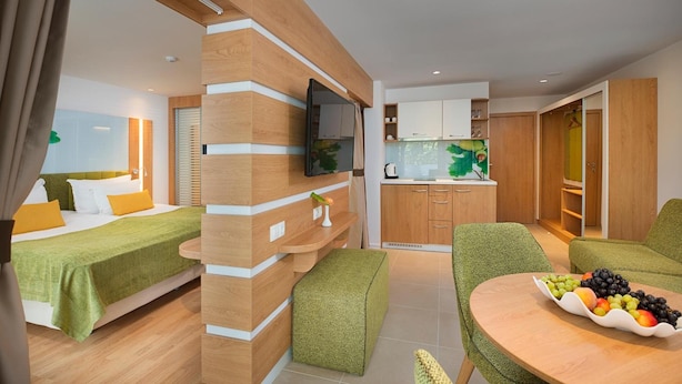 A modern hotel room with a bed, a small kitchenette, a dining area with a table and chairs, and a TV mounted on the wall.