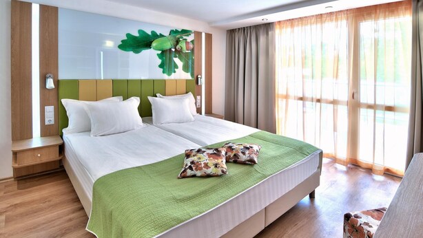 Ein Hotelzimmer mit einem großen Bett, einer grünen Decke und Blick ins Freie durch große Fenster.