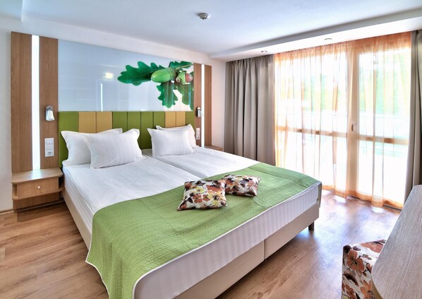 Ein Hotelzimmer mit einem großen Bett, einer grünen Decke und Blick ins Freie durch große Fenster.