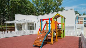 A playground with a slide and climbing area, surrounded by a white fence and a building with a white exterior.
