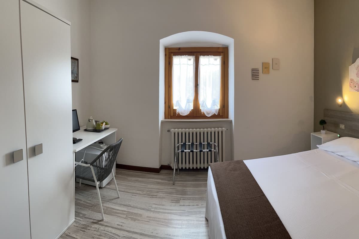 A hotel room with a bed, a desk, a radiator, and a window with curtains.