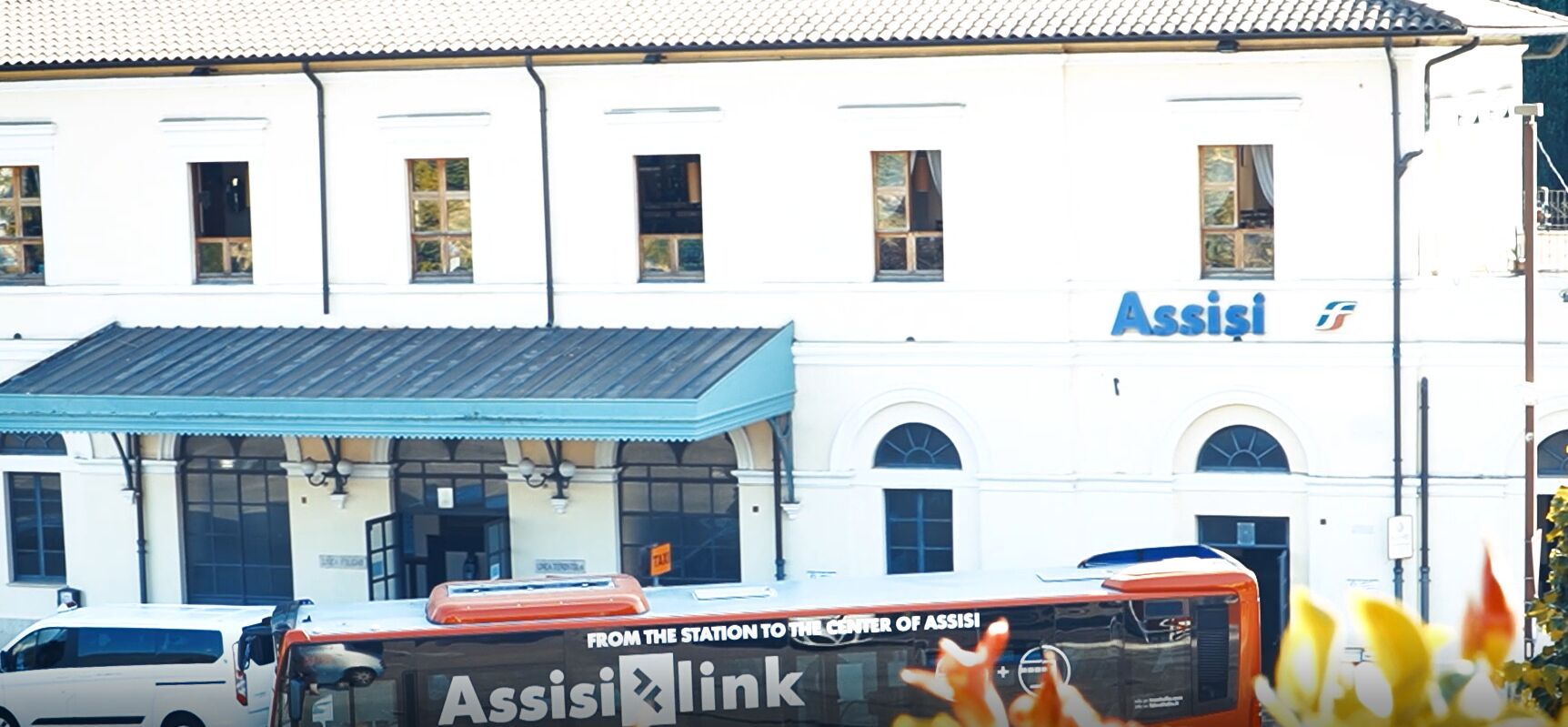Un bâtiment sur lequel on peut lire l’inscription « Assise », et un autobus sur lequel est écrit « Liaison Assise ».