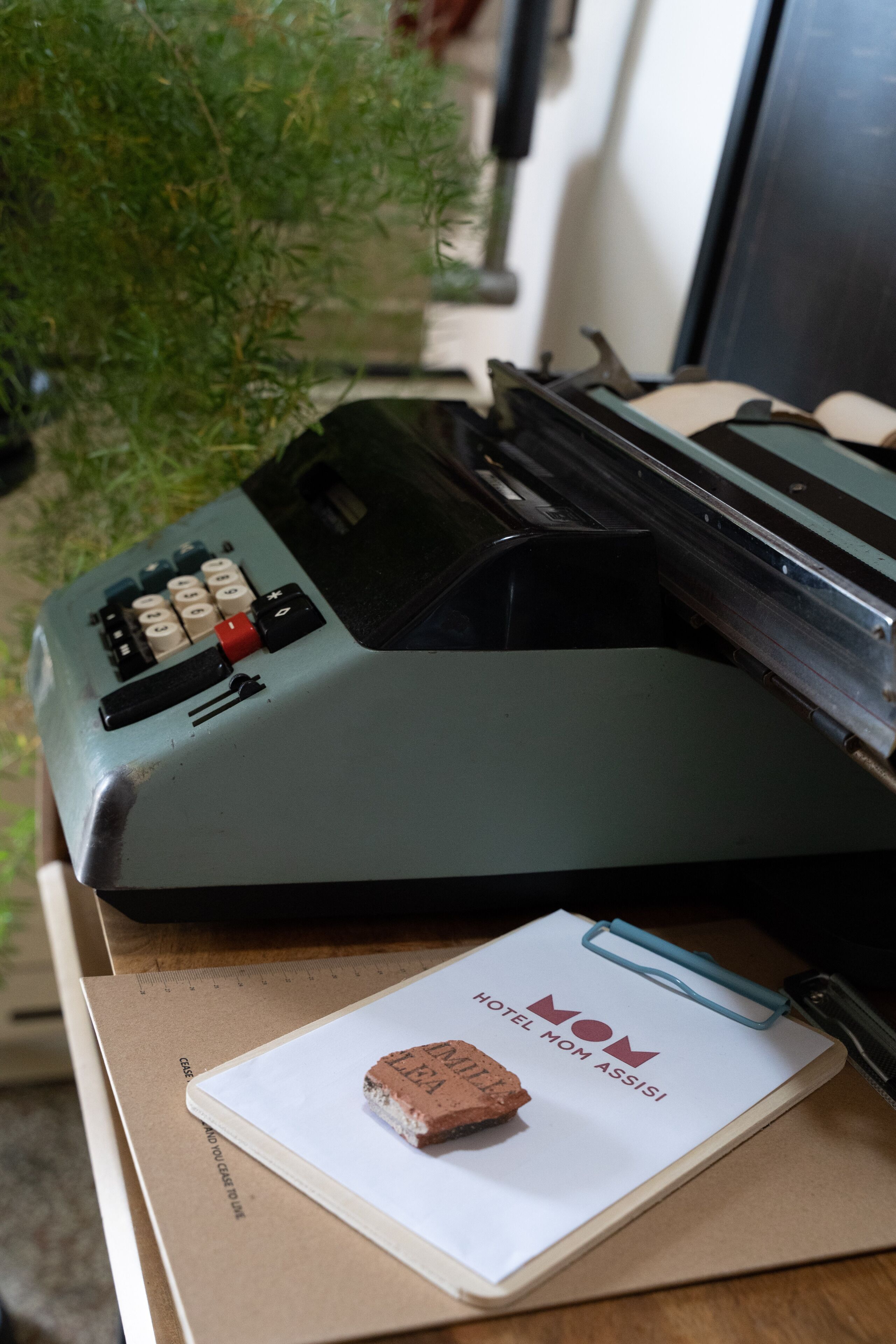 Una máquina de escribir antigua sobre un escritorio, junto a un cuaderno con la etiqueta "MOM HOTEL" y un ladrillo pequeño.