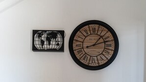 A living room with a brown leather sofa, a round wall clock, a small table with a vase, and a globe artwork on the wall. - Hotel Mom Assisi (Assisi)
