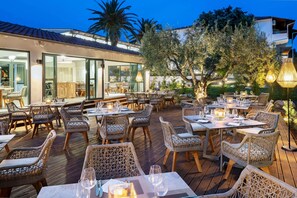 An outdoor dining area with wicker furniture and tables set for dining, surrounded by greenery and a building with large windows. - Philoxenia Hotel (Polygyros)