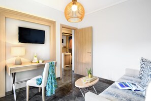 A modern hotel room with a flat-screen TV, a desk with a lamp, a chair, a sofa, a small table with a plant, and a magazine. - Philoxenia Hotel (Polygyros)