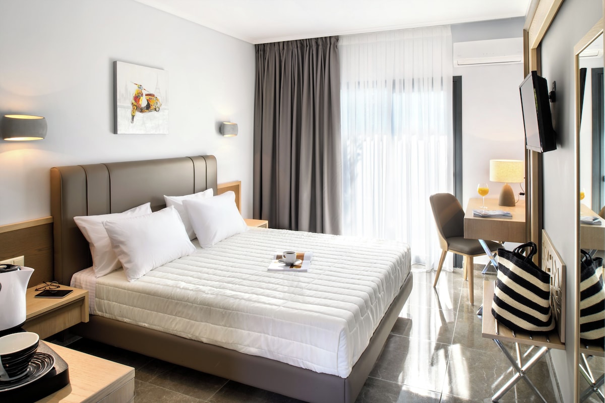 Habitació d'hotel amb un llit gran, una taula de treball, una cadira, un televisor i una finestra amb cortines.