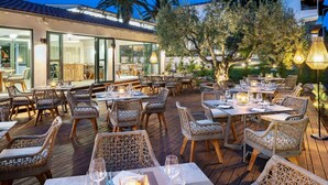 An outdoor dining area with wicker furniture and tables set for dining, surrounded by greenery and a building with large windows. - Philoxenia Hotel (Polygyros)