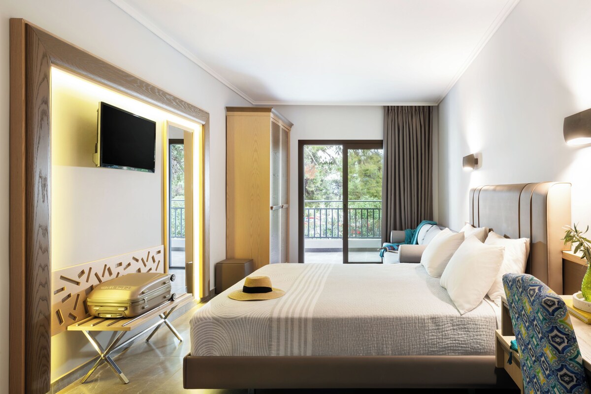 A hotel room with a large bed, a TV mounted on the wall, a suitcase on a bench, and a balcony with a view of trees.