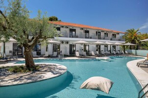 Área de piscina com espreguiçadeiras e guarda-sóis, uma árvore e um edifício com varandas.