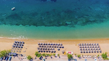 Vista aérea de una playa con filas de sombrillas y sillas de playa, un mar azul y despejado, y algunos barcos a lo lejos.