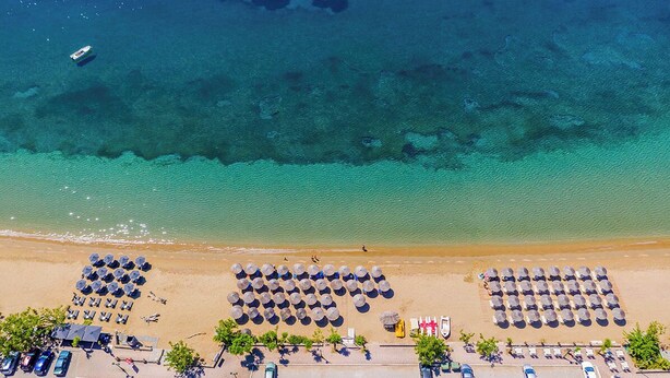 An aerial view of a beach with rows of sun umbrellas and lounge chairs, a clear blue sea, and a few boats in the distance. - Philoxenia Hotel (Polygyros)