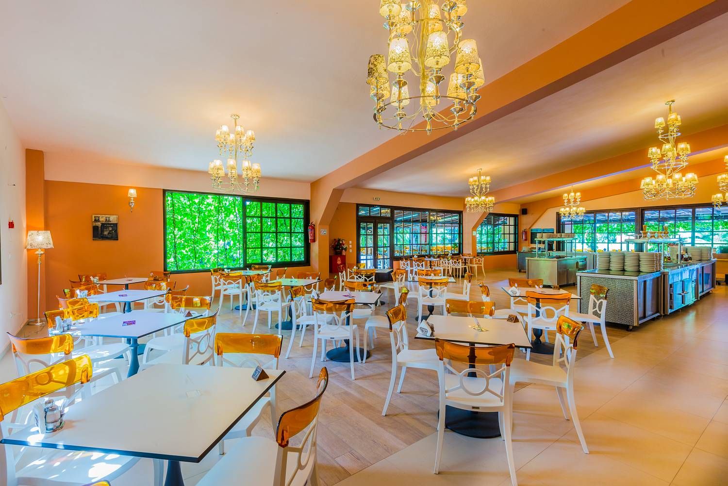 A spacious dining area with multiple tables and chairs, large windows with a view of greenery, and a kitchen area in the background.
