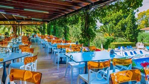 An outdoor dining area with tables and chairs, surrounded by lush greenery and a pool area. - Philoxenia Hotel (Polygyros)