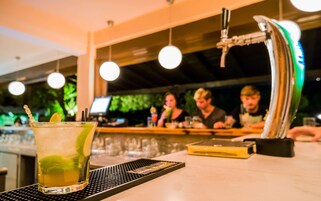 Uma área de bar com uma bebida no balcão, um caixa registradora e um fundo desfocado com pessoas.
