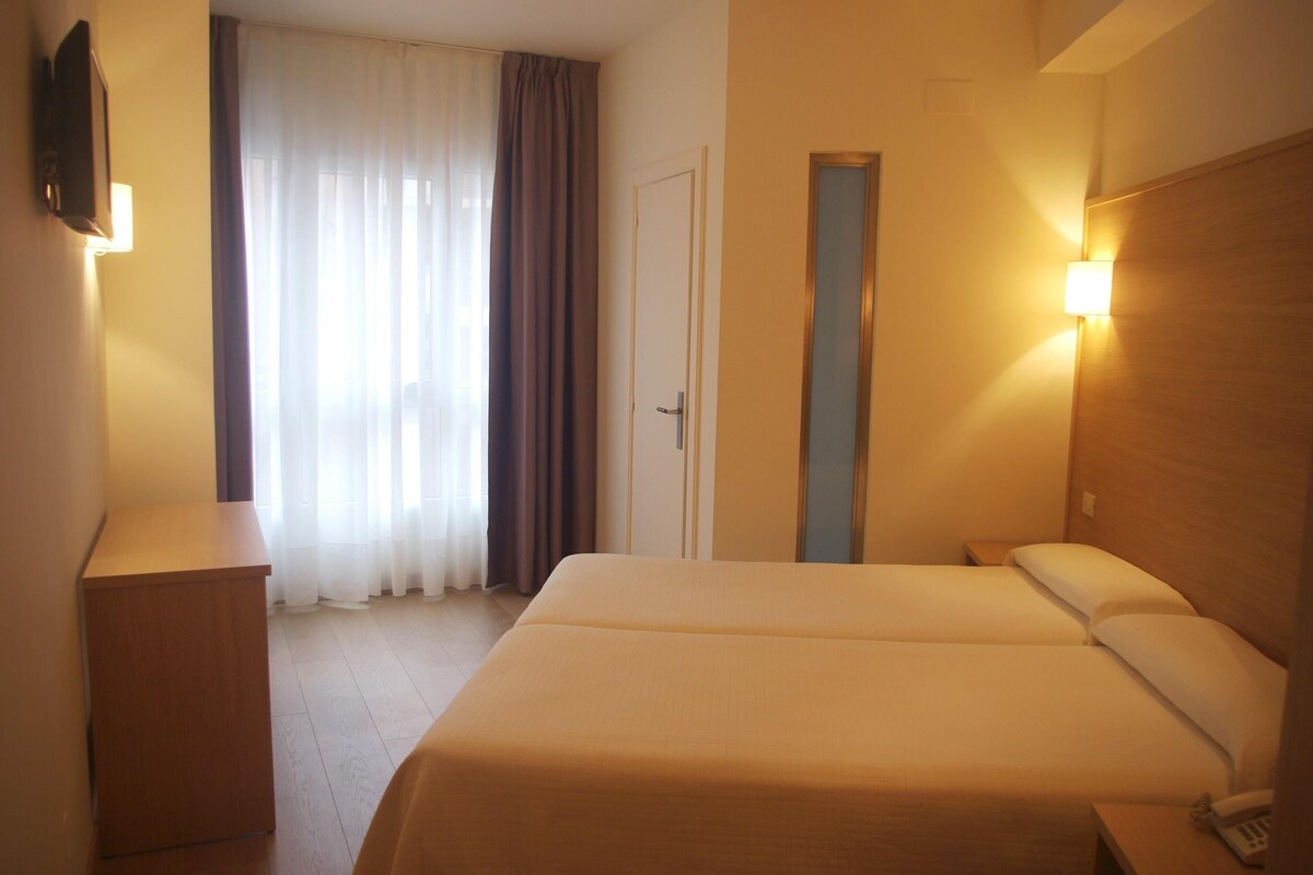 Een hotelkamer met een bed, nachtkastje, lamp en een deur.
